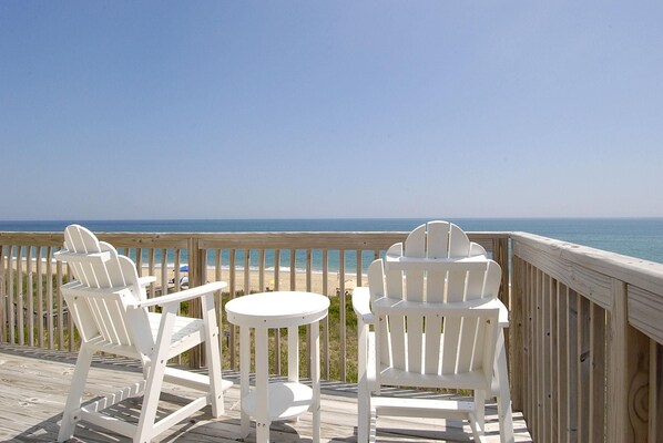 A wooden deck with white Adirondack chairs and a small round table, overlooking a beach and ocean.