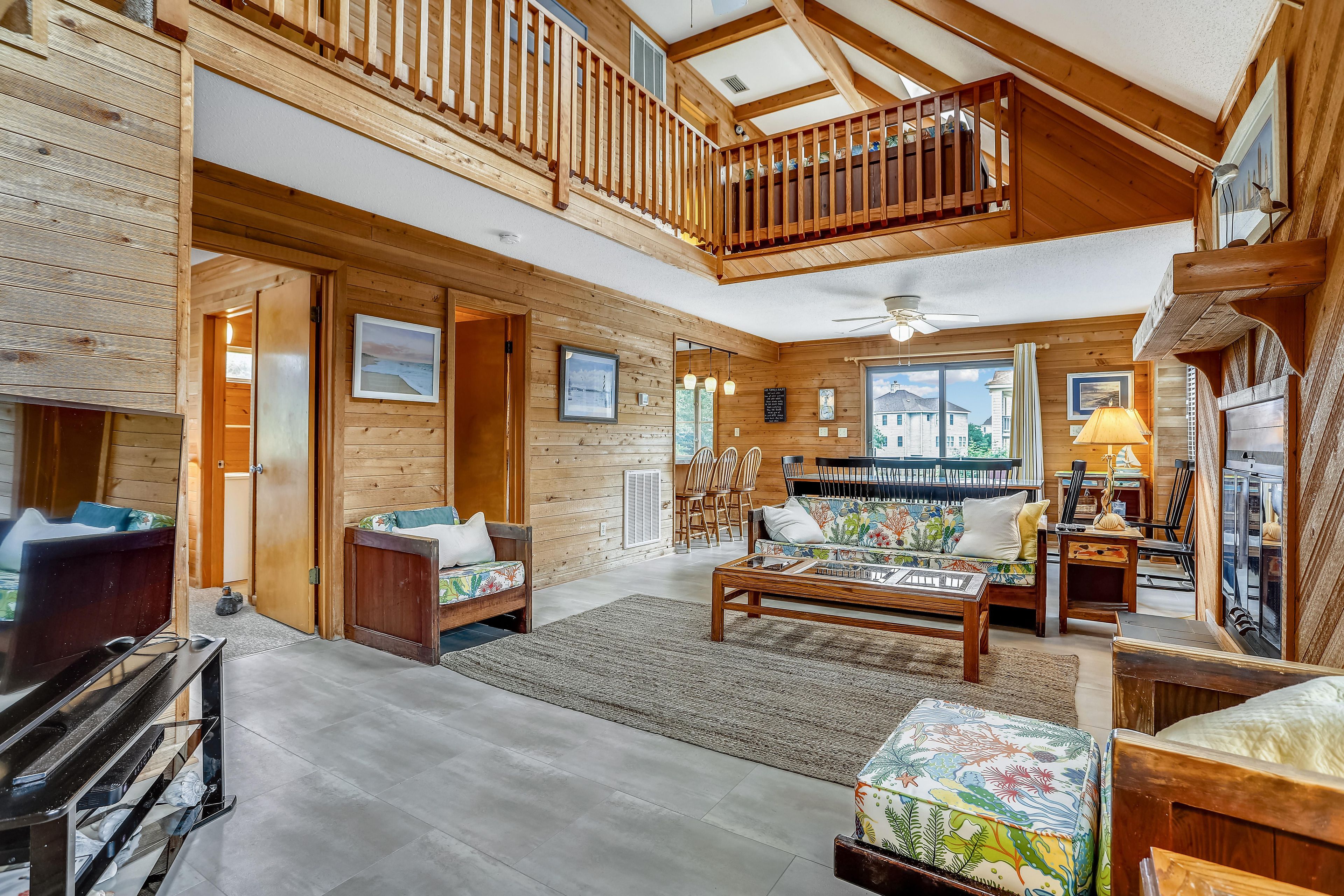 A spacious living area with a high ceiling, wooden beams, a balcony, and a dining area with chairs and a table.