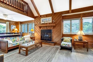 A cozy living room with a fireplace, wooden walls, and a view of the outdoors. - Timberland -- 4 BR Oceanside home in Southern Duck (Duck)