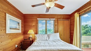 A wooden-paneled bedroom with a bed, a ceiling fan, a window with blinds, a nightstand with a lamp, and a framed picture on the wall.