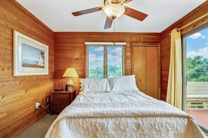 A wooden-paneled bedroom with a bed, a ceiling fan, a window with blinds, a nightstand with a lamp, and a framed picture on the wall. - Timberland -- 4 BR Oceanside home in Southern Duck (Duck)