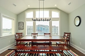 Uma sala de jantar com uma grande mesa de madeira, bancos e um candelabro. Há janelas com persianas, um relógio na parede e uma frase motivacional na parede.
