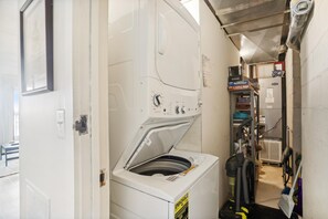A laundry room with a front-loading washing machine, a dryer, and a vacuum cleaner. - The Oceans Unit 301 -- Oceanfront Condo with Direct Access to Pool and Beach (Nags Head)
