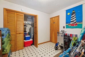 A room with a checkered floor, a closed wooden door, a painting of a lighthouse, and a wheelchair.