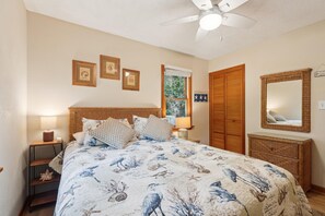 A bedroom with a bed, a ceiling fan, a window with blinds, a wooden wardrobe, and a wall-mounted mirror.