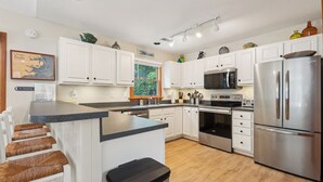 A modern kitchen with white cabinets, stainless steel appliances, a central island with stools, and a view of the outdoors. - Leeward Inn-- Beautifully Decorated Home with Pool, Hot Tub and More! (Southern Shores)