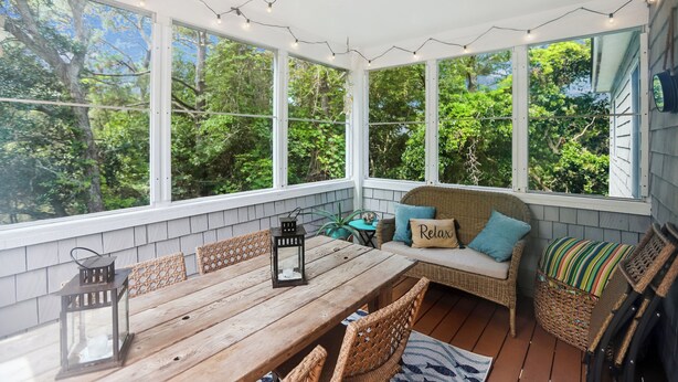 A sunroom with a wooden table, wicker chairs, and a cushioned seating area with a 'Relax' pillow. - Leeward Inn-- Beautifully Decorated Home with Pool, Hot Tub and More! (Southern Shores)