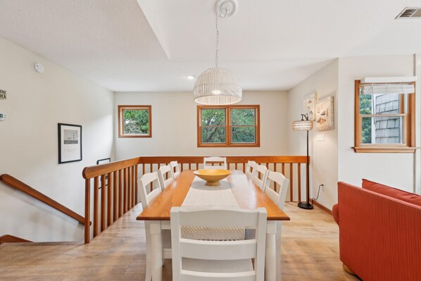 A dining area with a wooden table, white chairs, a hanging light fixture, and a view of the outdoors through windows.
