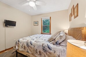 Une chambre avec un lit, un ventilateur de plafond, une fenêtre donnant sur un paysage verdoyant, un téléviseur fixé au mur et des tableaux encadrés accrochés au mur.