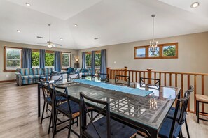 Uma sala de jantar ampla com mesa de vidro, cadeiras estofadas azuis e vista para o exterior através de grandes janelas.