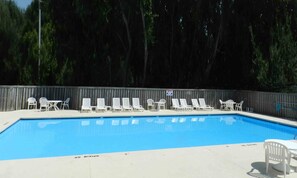 Une piscine entourée d’une clôture en bois et d’arbres, avec des chaises longues et des tables à proximité.