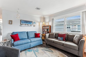 A living room with two sofas, a bookshelf, a painting, and a window with a view of the outside. - Blankenship Cottage -- Pet Friendly with Sunroom and Just 2 blocks to beach (Nags Head)