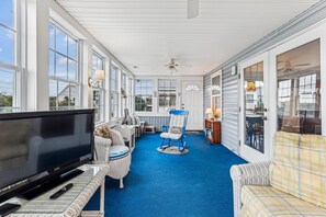 A sunroom with a large TV, wicker furniture, and a blue carpet. - Blankenship Cottage -- Pet Friendly with Sunroom and Just 2 blocks to beach (Nags Head)