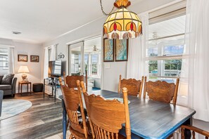 A dining area with a wooden table and chairs, a chandelier, and large windows.