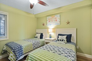 Ein Zimmer mit zwei Betten, jeweils mit gestreifter Bettdecke und passendem Kissen, einem Deckenventilator, einem an der Wand befestigten Schild und einem Fenster mit Jalousien.