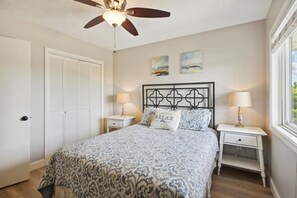 A bedroom with a bed, two nightstands, a ceiling fan, a door, and two wall-mounted paintings. - Tidewater Star -- Updated 2nd Floor Condo with Ocean Views and Pool (Duck)