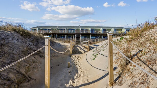 Ein Strandhotel mit einem Sandweg, der zum Eingang führt, Holzgeländern und blauem Himmel.