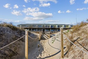 A beachfront hotel with a sandy pathway leading to the entrance, wooden railings, and a clear sky. - Seawalk Unit 8 -- Oceanfront Townhouse with Pool Access (Kill Devil Hills)
