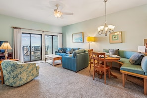 A spacious living room with a large sofa, a coffee table, and a dining area with chairs and a table. There is a ceiling fan, a chandelier, and a view through the glass doors. - Seawalk Unit 8 -- Oceanfront Townhouse with Pool Access (Kill Devil Hills)
