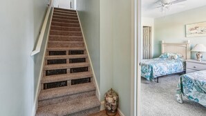 A carpeted staircase with a handrail, leading to a bedroom with two beds, a ceiling fan, and a wall-mounted light fixture. - Seawalk Unit 8 -- Oceanfront Townhouse with Pool Access (Kill Devil Hills)