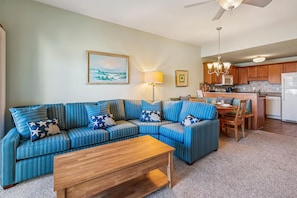 A living room with a sectional sofa, a wooden coffee table, a dining area with a table and chairs, and a kitchen with wooden cabinets and appliances. - Seawalk Unit 8 -- Oceanfront Townhouse with Pool Access (Kill Devil Hills)