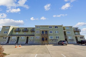 A multi-story residential building with a central entrance, balconies, and a parking area with a few vehicles. - Seawalk Unit 8 -- Oceanfront Townhouse with Pool Access (Kill Devil Hills)