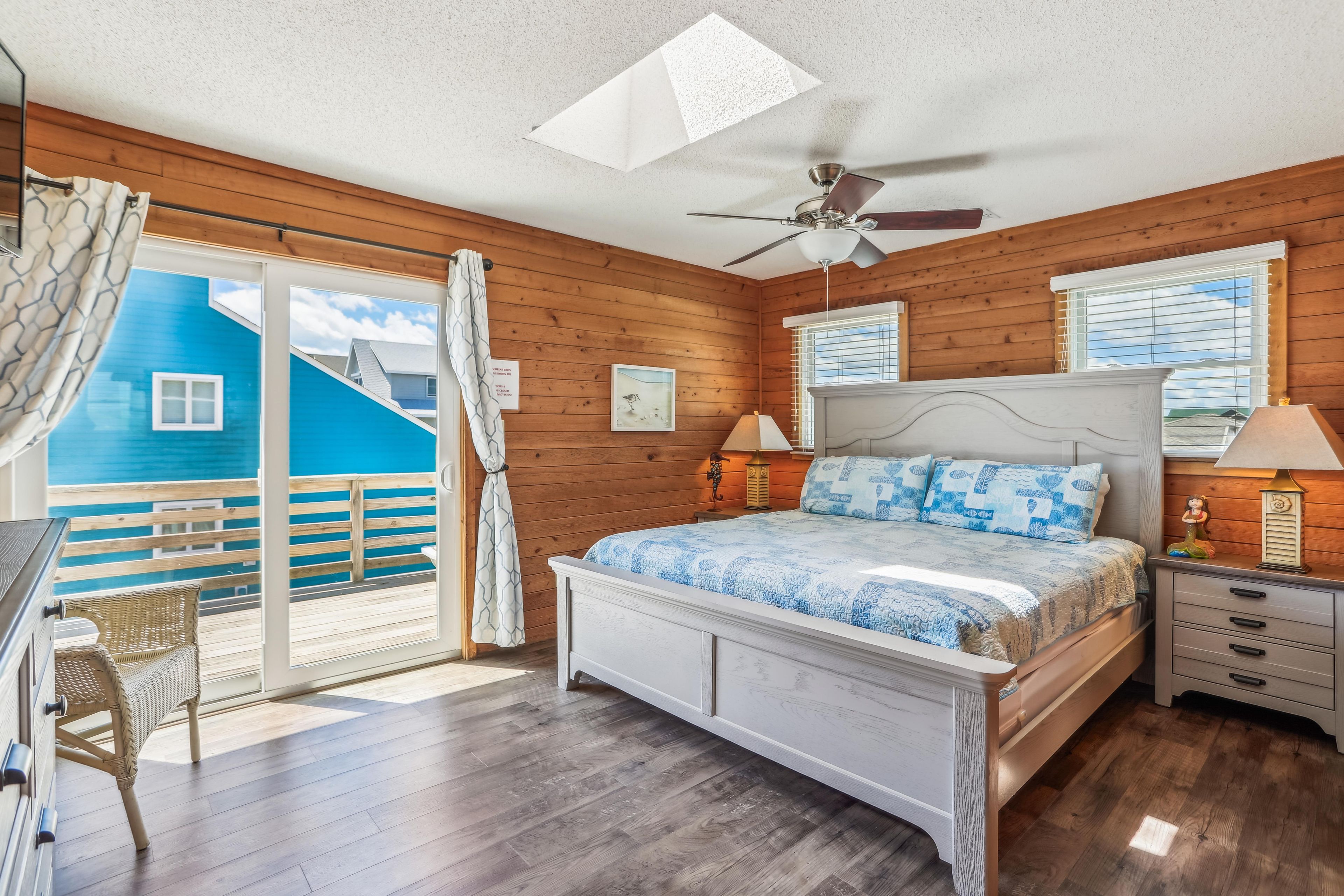 A bedroom with a bed, nightstands, a ceiling fan, a window with blinds, and a view of a blue building outside.