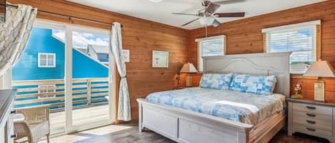 A bedroom with a bed, nightstands, a ceiling fan, a window with blinds, and a view of a blue building outside.