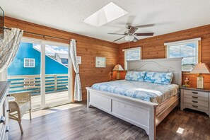 A bedroom with a bed, nightstands, a ceiling fan, a window with blinds, and a view of a blue building outside. - Freebird -- Pet Friendly Oceanfront in South Nags Head (Nags Head)