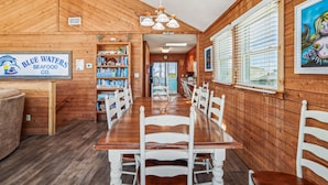 Un intérieur en bois avec une longue table à manger, des chaises blanches, une bibliothèque et un panneau indiquant « Blue Waters Seafood Co. ».