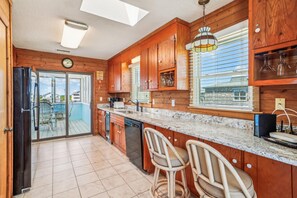 A kitchen with wooden cabinets, a dishwasher, a sink, and a bar area with chairs. - Freebird -- Pet Friendly Oceanfront in South Nags Head (Nags Head)