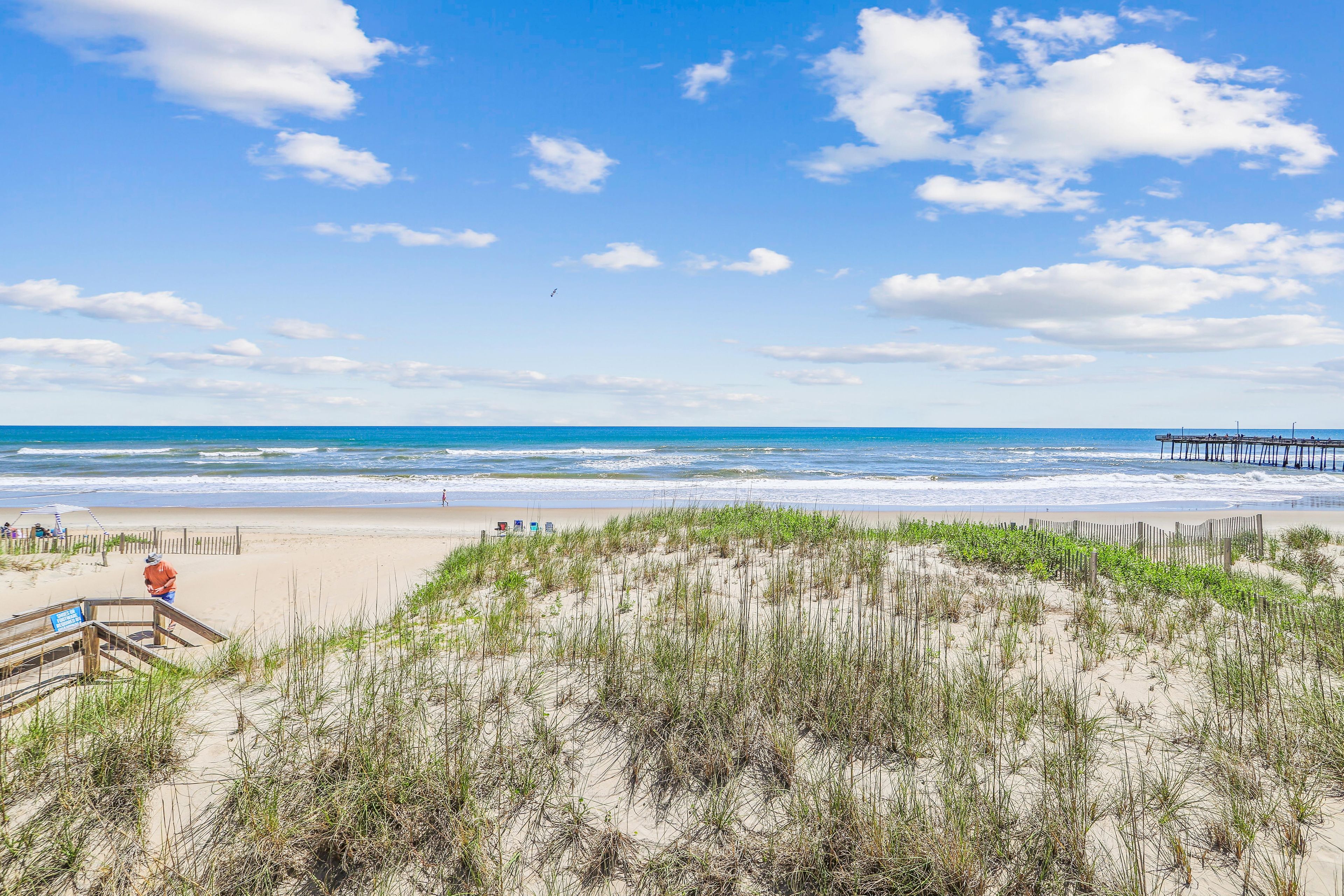 Ein Strand mit einer hölzernen Steg, der sich ins Meer erstreckt, einem sandigen Bereich mit Gras und einer Treppe, die zum Strand hinabführt.