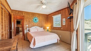 Une chambre à coucher avec un lit, un ventilateur de plafond, des murs en bois, une fenêtre avec des stores et une vue sur l’extérieur.