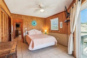 Un dormitorio con cama, ventilador de techo, paredes de madera, ventana con persianas y vistas al exterior.