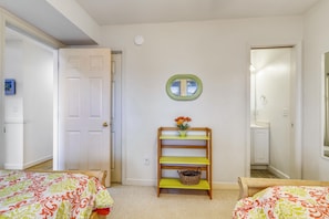 Une chambre avec deux lits, un banc en bois avec un panier, un miroir fixé au mur et une porte donnant sur une autre pièce.