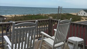A wooden deck with two rocking chairs and a small stool, overlooking a beach with a fence and a white building in the distance. - Miller Cottage -- Semi OF Home in Kitty Hawk with Hot Tub (Kitty Hawk)