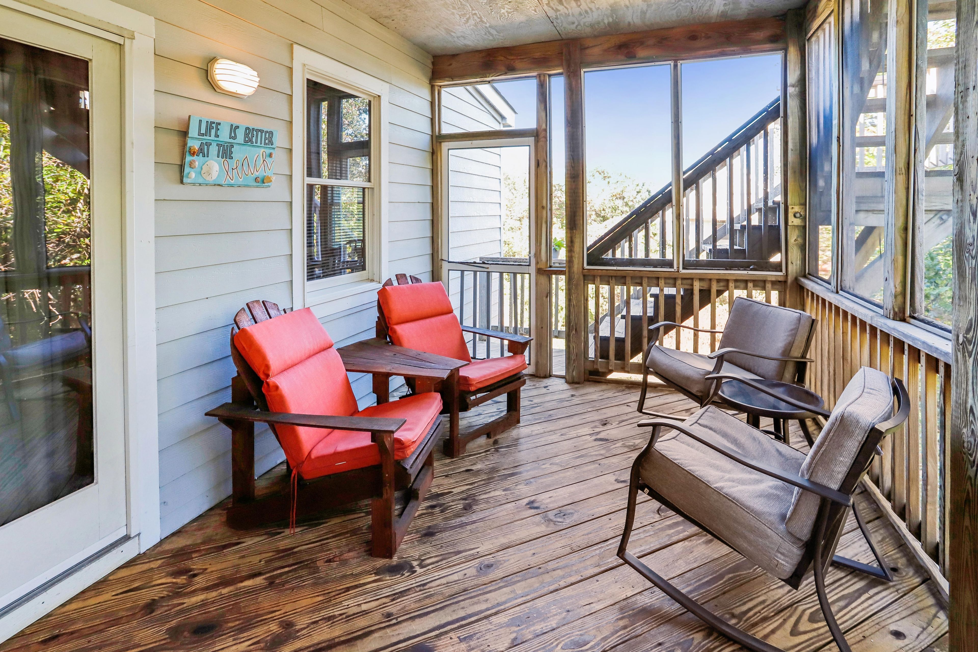 A wooden deck with outdoor seating, including two red chairs and a bench, and a sign that reads 'Life is better at the beach.'