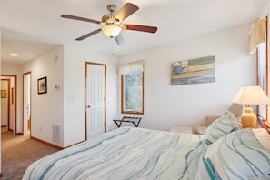 Un dormitorio con cama, ventilador de techo, ventana con persianas, una obra de arte en la pared y una lámpara de noche.
