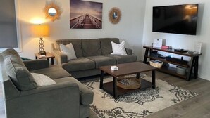 A living room with a sofa, armchair, coffee table, TV, and a small side table with a lamp. - Flamingo -- Pet Friendly Home with Private Pool and Gameroom (Nags Head)