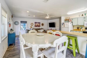 A dining area with a white table and chairs, a blue side table, a green stool, and a kitchen area with light green cabinets and a white refrigerator. - Flamingo -- Pet Friendly Home with Private Pool and Gameroom (Nags Head)