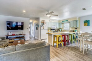 A modern living room with a grey sofa, a wooden coffee table, and a flat-screen TV. A kitchen with light green cabinets and a wooden island with stools is visible. - Flamingo -- Pet Friendly Home with Private Pool and Gameroom (Nags Head)