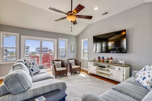 A spacious living room with a ceiling fan, large windows, a flat-screen TV, and a view of the beach. - Paws in the Sand -- Gorgeous Pet Friendly Home with Pool (Kitty Hawk)