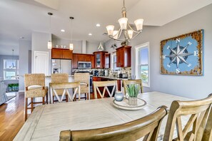 A modern kitchen with wooden cabinets, a central island, and a dining area with a table and chairs. - Paws in the Sand -- Gorgeous Pet Friendly Home with Pool (Kitty Hawk)