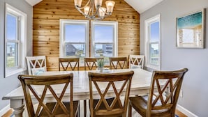 A dining area with a wooden table and chairs, large windows, and a chandelier. - Paws in the Sand -- Gorgeous Pet Friendly Home with Pool (Kitty Hawk)