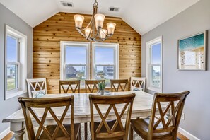 A dining area with a wooden table and chairs, large windows, and a chandelier. - Paws in the Sand -- Gorgeous Pet Friendly Home with Pool (Kitty Hawk)