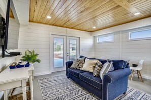 Une pièce avec un plafond en bois, un canapé bleu orné de coussins à motifs, un bureau blanc avec une plante et un tapis à motifs.