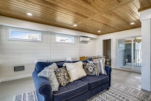 Une pièce avec un plafond en bois, un canapé bleu orné de coussins à motifs, une petite table et une fenêtre donnant sur un immeuble.
