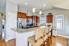 A kitchen with a granite countertop, stainless steel appliances, wooden cabinets, and a bar area with woven chairs. - Paws in the Sand -- Gorgeous Pet Friendly Home with Pool (Kitty Hawk)