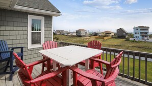 A deck with a white table and red chairs, overlooking a beach with houses in the background. - Paws in the Sand -- Gorgeous Pet Friendly Home with Pool (Kitty Hawk)