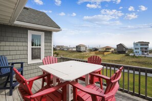 A deck with a white table and red chairs, overlooking a beach with houses in the background. - Paws in the Sand -- Gorgeous Pet Friendly Home with Pool (Kitty Hawk)